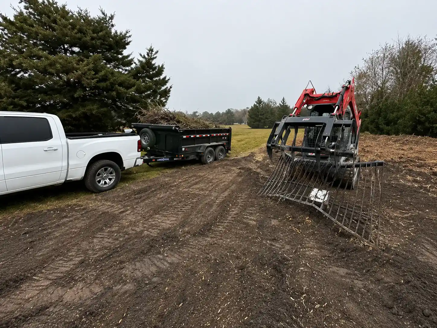 Dump trailer loaded with brush beside a skid steer with a grapple rake on freshly mulched ground